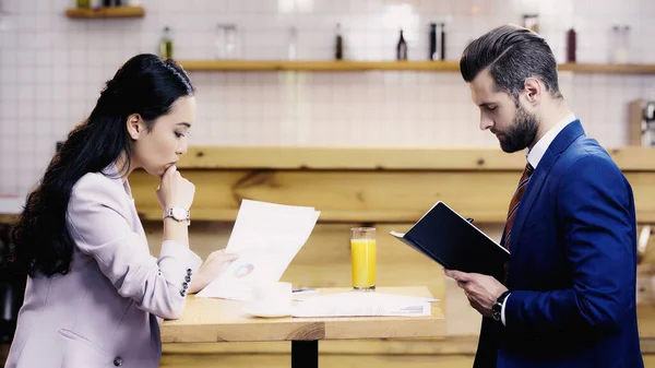 Vue de côté de coûteux asiatique femme d'affaires regardant document près barbu homme d'affaires dans le café — Photo de stock