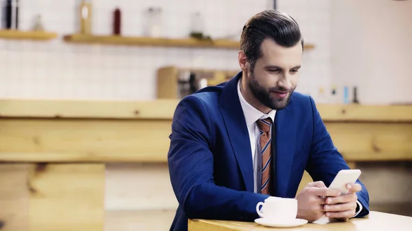 Hombre de negocios alegre en traje usando teléfono inteligente en la cafetería - foto de stock