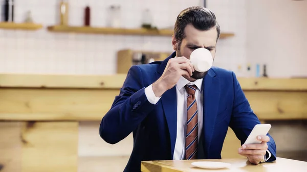 Bearded businessman in suit using smartphone and drinking coffee in cafe — Stock Photo