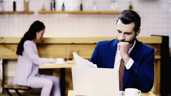 Hombre de negocios barbudo mirando los papeles cerca del ordenador portátil y la mujer de negocios borrosa en la cafetería - foto de stock