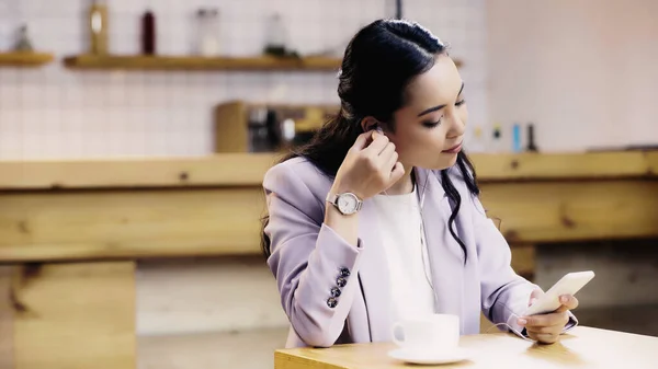 Jeune femme asiatique en costume écouter de la musique dans les écouteurs et en utilisant smartphone dans le café — Photo de stock