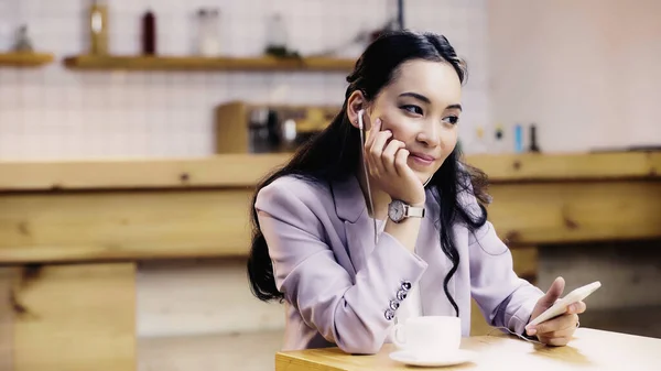 Mujer asiática complacida en traje escuchando música en auriculares y sosteniendo teléfono inteligente cerca de la taza de café en la cafetería - foto de stock