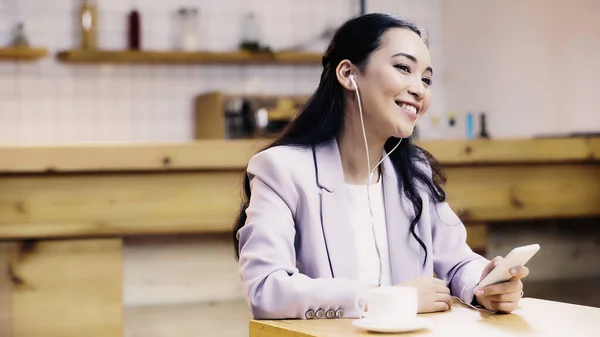 Щаслива азіатська жінка в костюмі слухає музику в навушниках і тримає мобільний телефон біля чашки в кафе — стокове фото