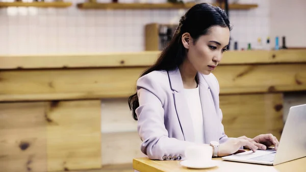 Focused and pretty asian businesswoman in suit typing on laptop while working remotely in cafe — Stock Photo