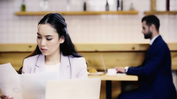 Seria asiático mujer de negocios mirando los papeles cerca borrosa hombre de negocios en fondo en café - foto de stock