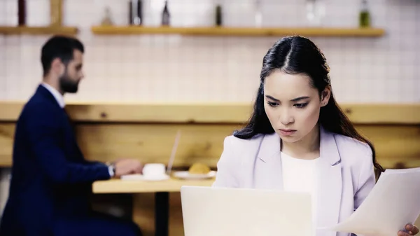 Серйозна азіатська бізнес-леді, дивлячись на документи поблизу розмитого бізнесмена на фоні кафе — стокове фото
