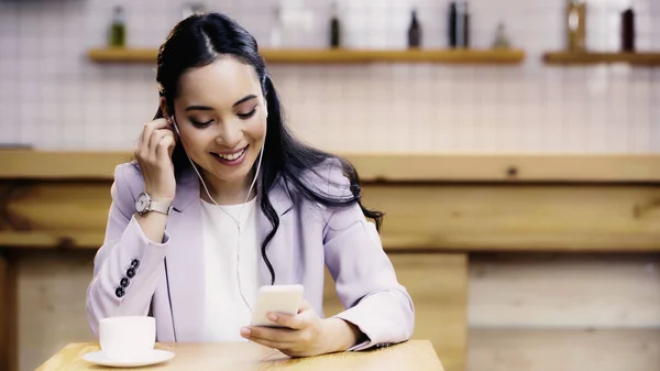 Feliz mulher asiática em terno ajustando fones de ouvido e segurando smartphone perto da xícara de café no café — Fotografia de Stock