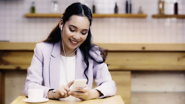 Mujer asiática complacida escuchando música en auriculares y sosteniendo teléfono inteligente cerca de la taza de café en la cafetería - foto de stock