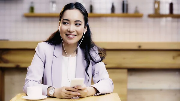 Feliz asiático mujer de negocios en traje escuchar música en auriculares cerca de taza de café en la cafetería - foto de stock