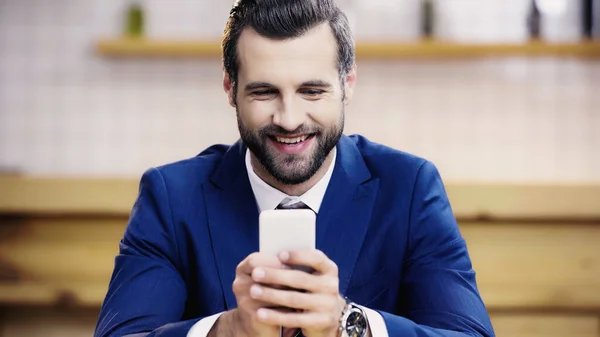 Bärtiger Geschäftsmann im Anzug lächelt, während er im Café per Handy Nachrichten verschickt — Stockfoto
