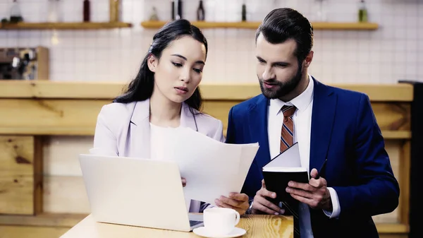 Barbudo empresario celebración notebook cerca asiático mujer de negocios trabajando remotamente en café - foto de stock