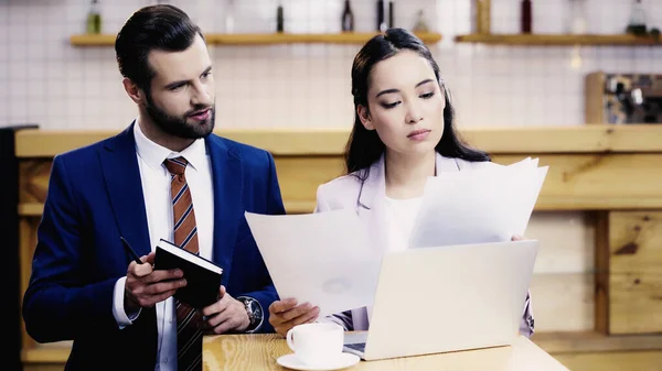 Barbudo hombre de negocios mirando documentos cerca asiático mujer de negocios trabajando remotamente en café - foto de stock