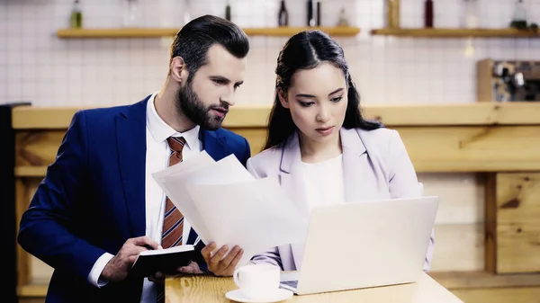 Bearded businessman holding notebook near asian businesswoman working remotely near laptop in cafe — Stock Photo