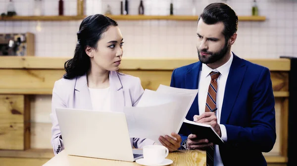 Barbu homme d'affaires écriture dans notebook près asiatique femme d'affaires travaillant à distance dans le café — Photo de stock