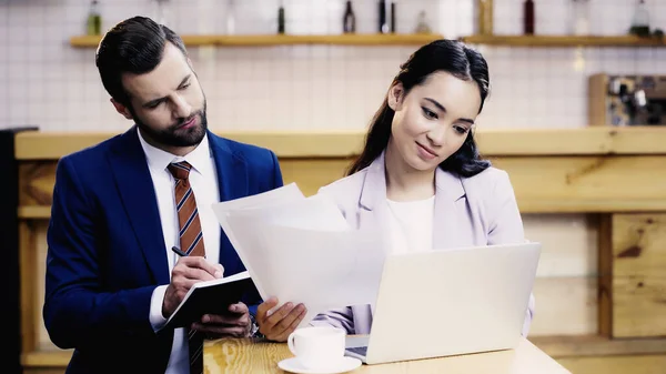 Bärtige Geschäftsfrau schreibt in Notizbuch in der Nähe erfreut asiatische Geschäftsfrau, die aus der Ferne in Café arbeitet — Stockfoto