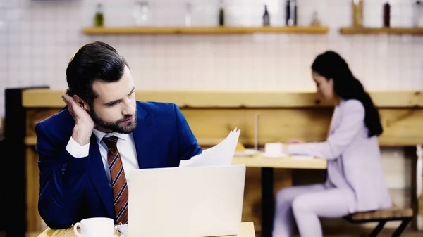 Бородатий бізнесмен дивиться на документи біля ноутбука і чашки на столі в кафе — стокове фото