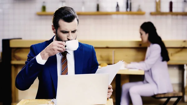 Hombre de negocios barbudo sosteniendo documentos cerca del ordenador portátil y beber café - foto de stock