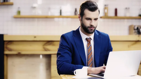 Bearded businessman in suit using laptop near cup in cafe — Stock Photo