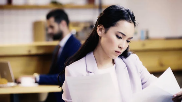 Concentrated asian businesswoman looking at documents near blurred businessman on background in cafe — Stock Photo