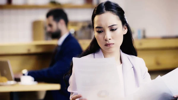 Fokussierte asiatische Geschäftsfrau schaut sich Dokumente in der Nähe verschwommener Geschäftsmann an — Stockfoto