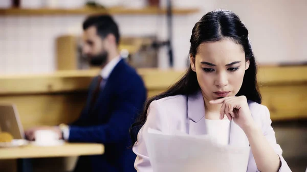 Focused asian businesswoman looking at documents near blurred businessman on background in cafe — Stock Photo