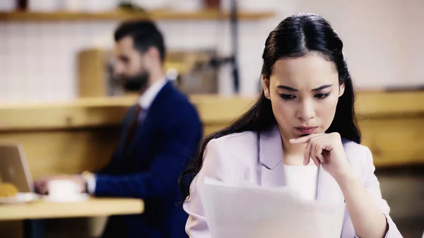 Focalizzato asiatico businesswoman guardando documenti vicino offuscata businessman su sfondo in caffè — Foto stock