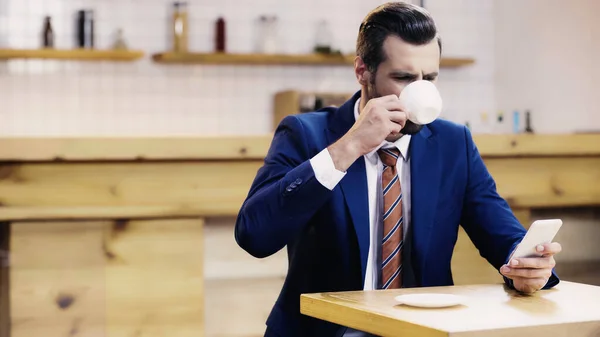 Homem de negócios barbudo em terno beber café e usar smartphone no café — Fotografia de Stock
