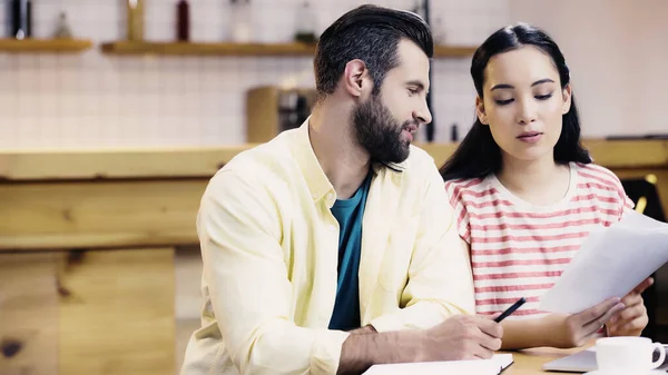 Interracial students looking at conspectus near cup on table in cafe — Stock Photo