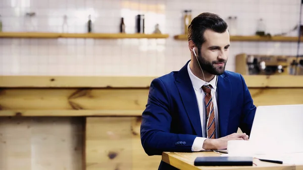 Усміхаючись бізнесмен у навушниках за допомогою ноутбука біля чашки в кафе. — стокове фото