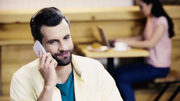 Бородатий чоловік говорить по смартфону біля розмитої жінки в кафе — Stock Photo