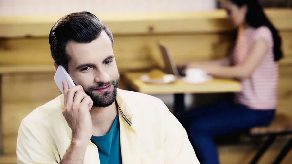 Bearded man talking on smartphone near blurred woman in cafe — Stock Photo