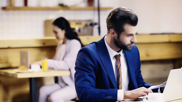 Sorprendido hombre de negocios mirando los documentos cerca de la computadora portátil y la mujer borrosa en la cafetería - foto de stock