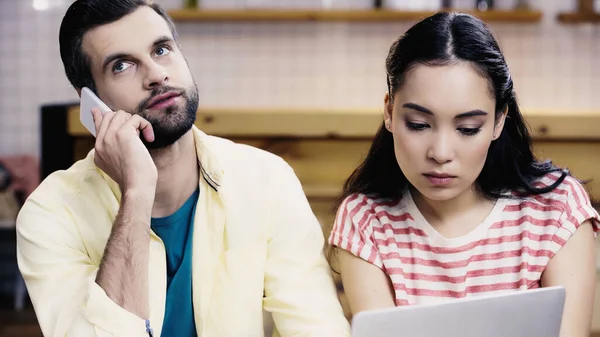 Азиатский студент смотрит на ноутбук рядом бородатый мужчина разговаривает на смартфоне в кафе — стоковое фото