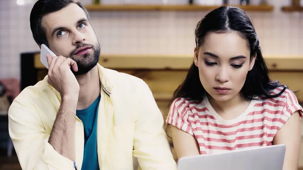 Asiatischer Student schaut in Café auf Laptop neben bärtigem Mann, der mit Smartphone spricht — Stockfoto