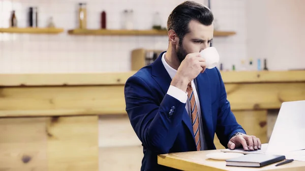Бородатий бізнесмен в костюмі п'є каву при використанні ноутбука в кафе — стокове фото