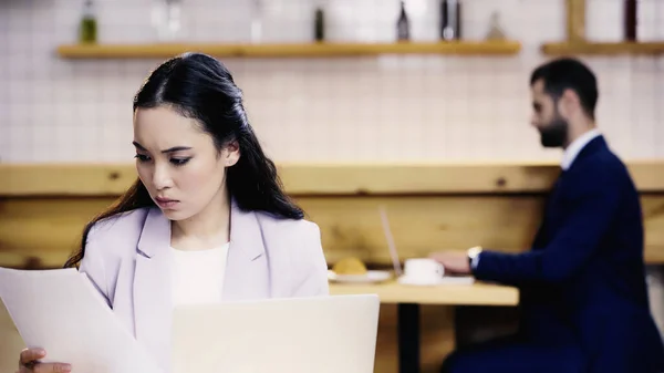 Young asian businesswoman looking at documents near blurred businessman in cafe — Stock Photo