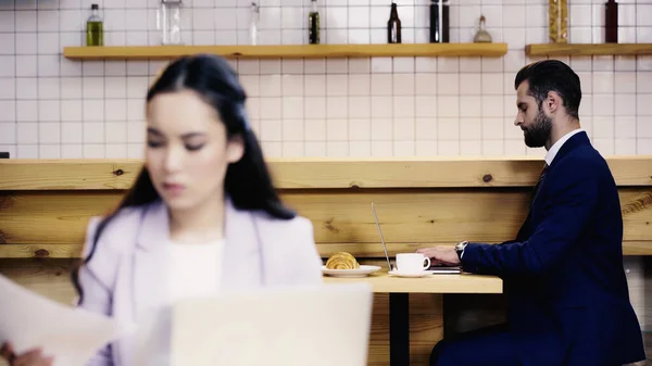 Hombre de negocios escribiendo en el ordenador portátil cerca borrosa mujer asiática con documentos en primer plano - foto de stock