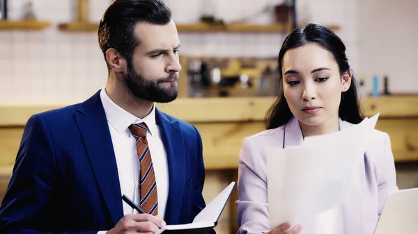 Bearded businessman writing in notebook near asian businesswoman with documents in cafe — Stock Photo