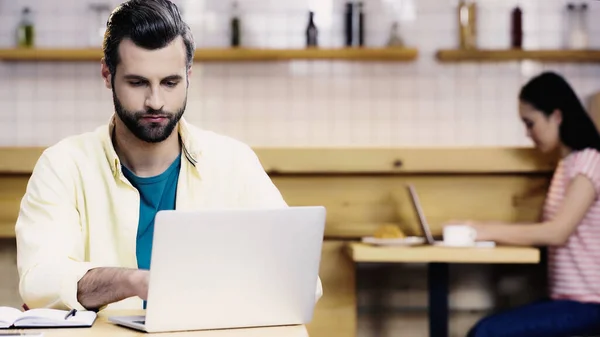 Bärtiger Mann mit Laptop neben Notizbuch auf Tisch und verschwommene Frau in Café — Stockfoto