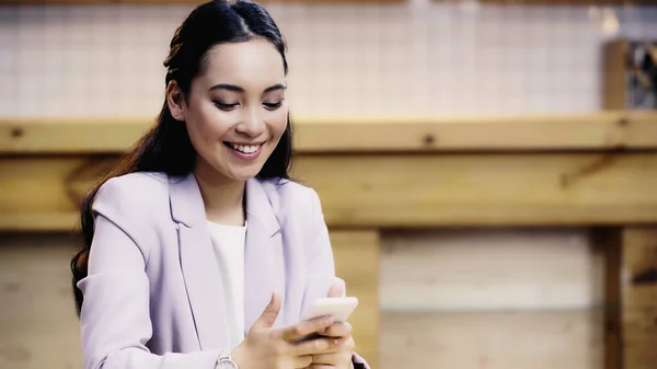 Pleased asian businesswoman in suit using smartphone in cafe — Stock Photo