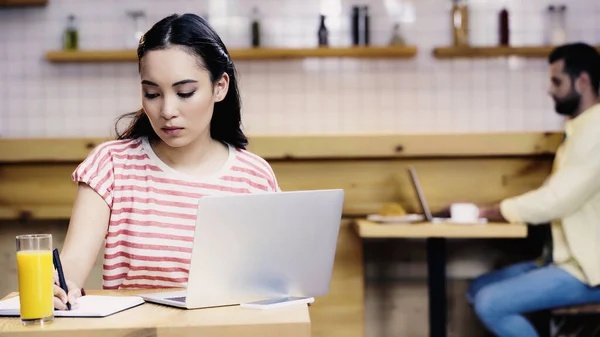 Asian woman in t-shirt writing in notebook near laptop and blurred man on background — Stock Photo