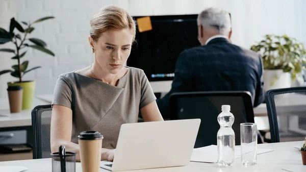 Donna d'affari che utilizza laptop vicino a caffè e acqua in ufficio — Foto stock