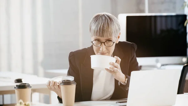 Mature businesswoman smelling takeaway lunch near laptop in office — Stock Photo