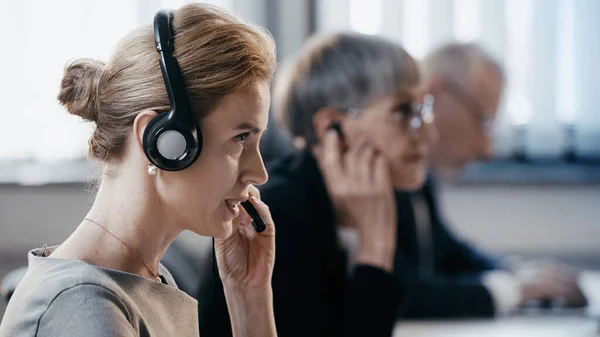 Vista lateral de la mujer de negocios que usa auriculares mientras trabaja cerca de colegas borrosos en la oficina - foto de stock