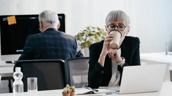 Femme d'affaires mature buvant du café pour aller près de l'ordinateur portable au bureau — Photo de stock