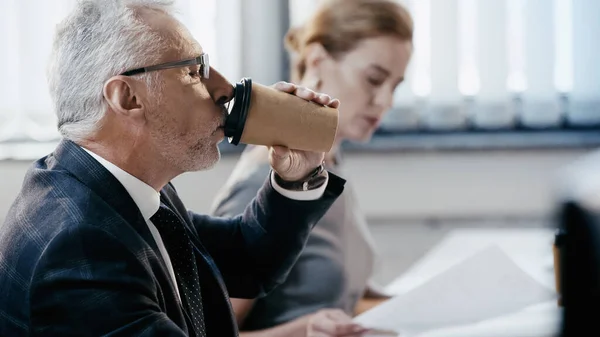 Side view of businessman drinking coffee to go near blurred colleague in office — Stock Photo