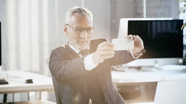 Зрелый бизнесмен делает селфи на смартфоне рядом с ноутбуком в офисе — стоковое фото