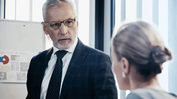 Reifer Geschäftsmann mit Brille blickt verschwommenen Kollegen nahe Flipchart im Büro an — Stockfoto