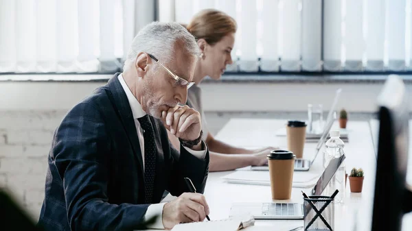 Side view of mature businessman writing on notebook near laptops in office — Stock Photo