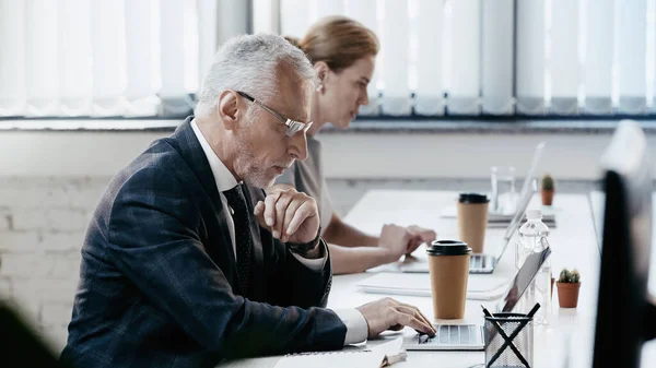 Боковой вид зрелого бизнесмена, использующего ноутбук рядом с кофе в офисе — стоковое фото